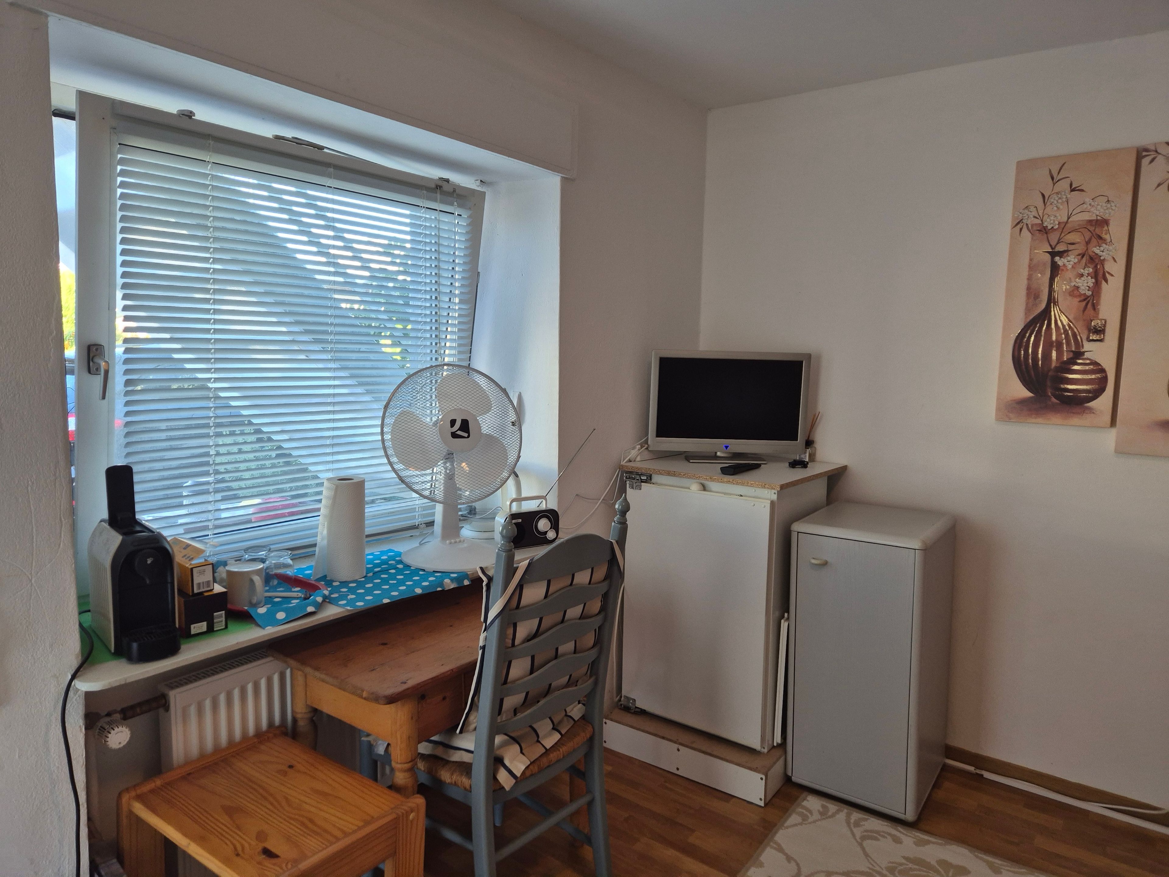 Ein gemütlicher, kleiner Wohnraum mit Schreibtisch, Stuhl, Heizkörper, kleinem Kühlschrank und einem Fenster mit Jalousien.