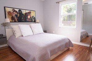 A bedroom with a bed, a nightstand, a lamp, a painting, and a window with a view of greenery. - Home with gorgeous backyard and pool in Fort Lauderdale (Fort Lauderdale)