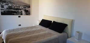 Ein Schlafzimmer mit einem Bett, einem Nachttisch, einer Wandlampe und einem Bilderrahmen mit einem Motiv einer Küstenstadt.