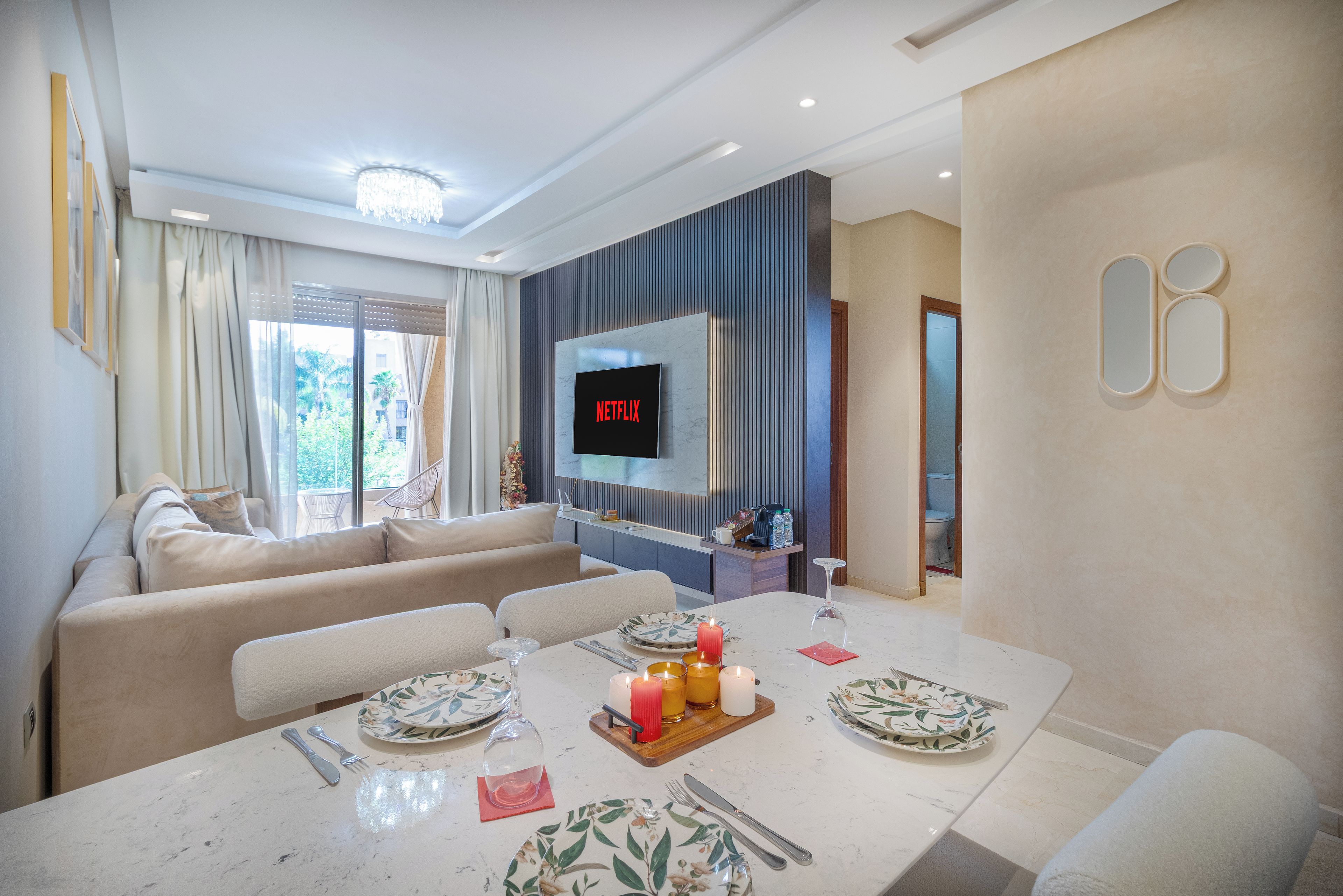 Una sala de estar moderna con un televisor de pantalla plana montado en la pared, un comedor con una mesa preparada para dos personas, y un sofá al fondo.