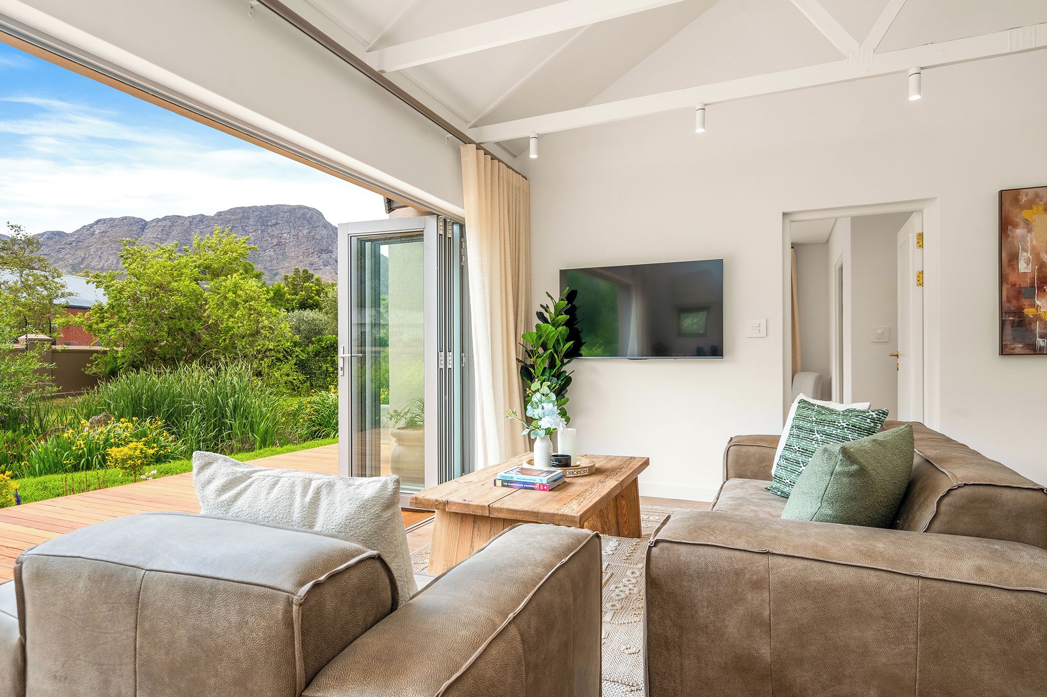 A modern living room with a sofa, a coffee table, and a TV. There is a large window with a view of greenery and mountains.