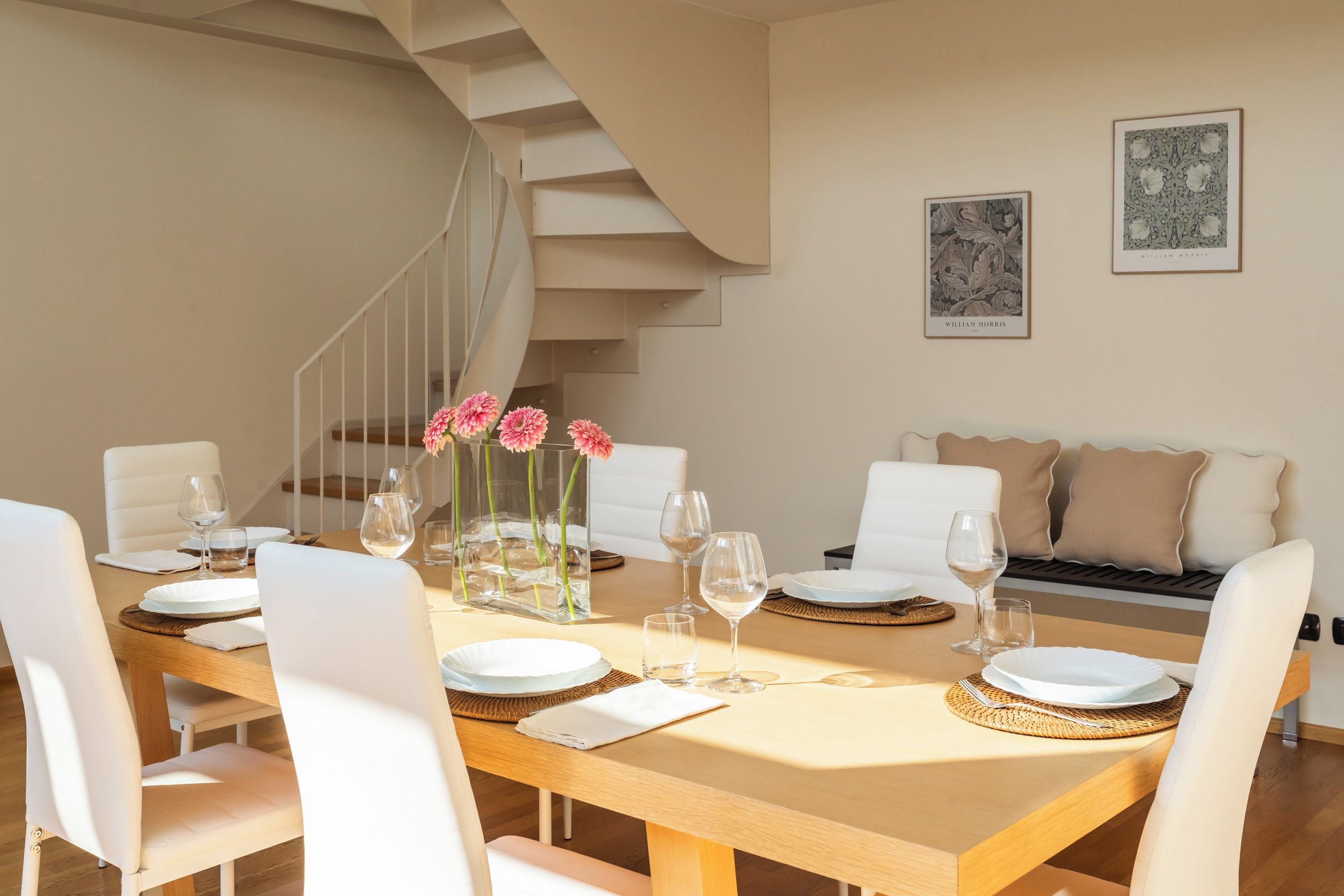 Un coin repas avec une longue table dressée pour six personnes, des chaises blanches et un vase rempli de fleurs roses.