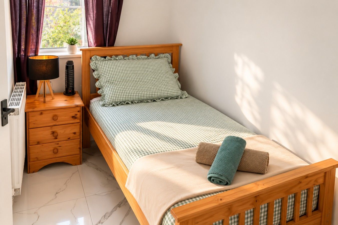 Ein Einzelbett mit karierter Bettdecke, ein Nachttisch aus Holz mit Schubladen, eine Lampe und ein gerolltes Handtuch auf dem Bett.