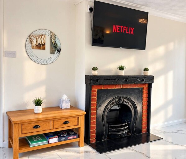 Un soggiorno con camino, una TV a parete che mostra 'NETFLIX', un tavolino di legno con libri e uno specchio decorativo.