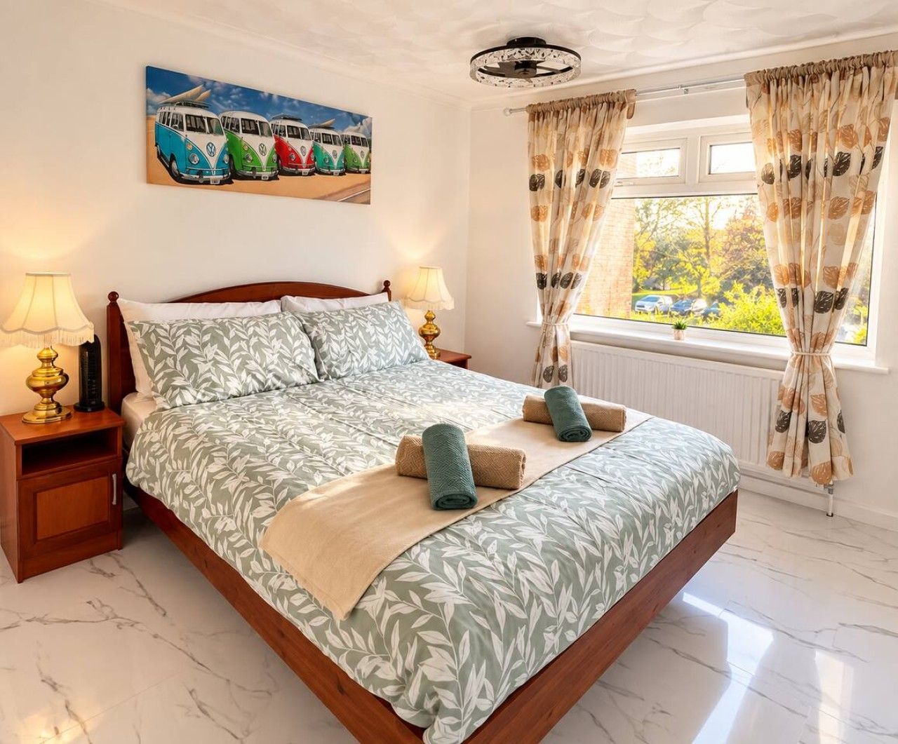 Uma cama bem arrumada com colcha estampada em folhagens, duas luminárias de cabeceira, um quadro com carros antigos e uma janela com cortinas.