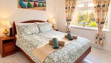 Uma cama bem arrumada com colcha estampada em folhagens, duas luminárias de cabeceira, um quadro com carros antigos e uma janela com cortinas.
