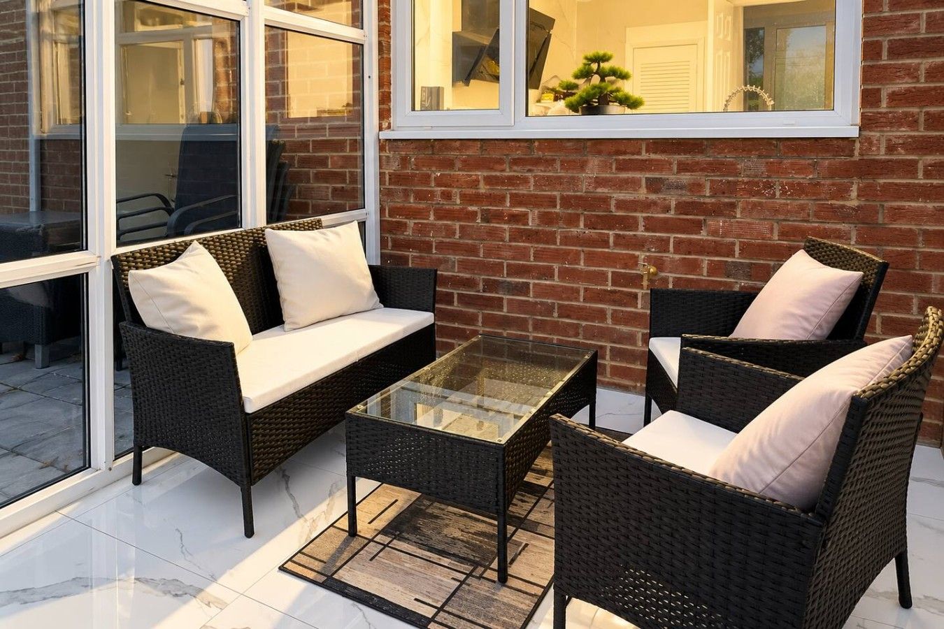 An outdoor seating area with wicker furniture and a glass table, adjacent to a brick wall and large windows.