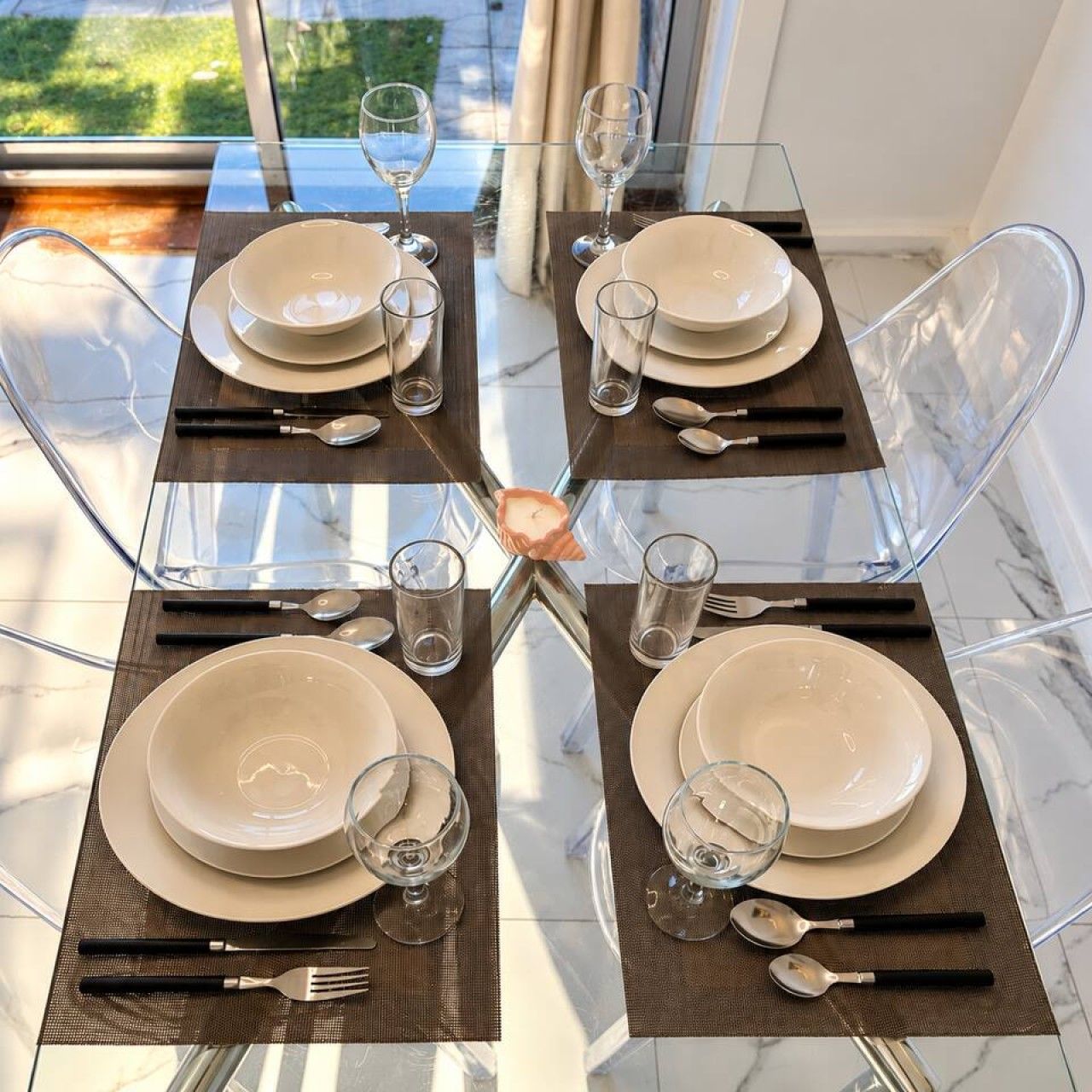 A dining table set for four with clear glassware and plates, positioned in a room with a view of greenery outside.