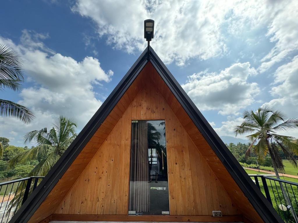 Una estructura de madera triangular con una puerta y una ventana pequeña, rodeada de palmeras y un cielo despejado.
