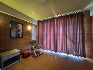 Una habitación con pared de ladrillo, un sillón acolchado, un pequeño otoman y una ventana con cortinas.