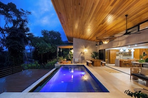 A modern outdoor pool area with a wooden deck, a lounge area with seating, and a kitchen visible through an open doorway. - Hidden Villa Oasis: Infinity Pool & Ocean Views near Dominical (Savegre de Aguirre)