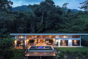 Una casa moderna de dos pisos amb una piscina, envoltada de vegetació exuberant i un fons de bosc.