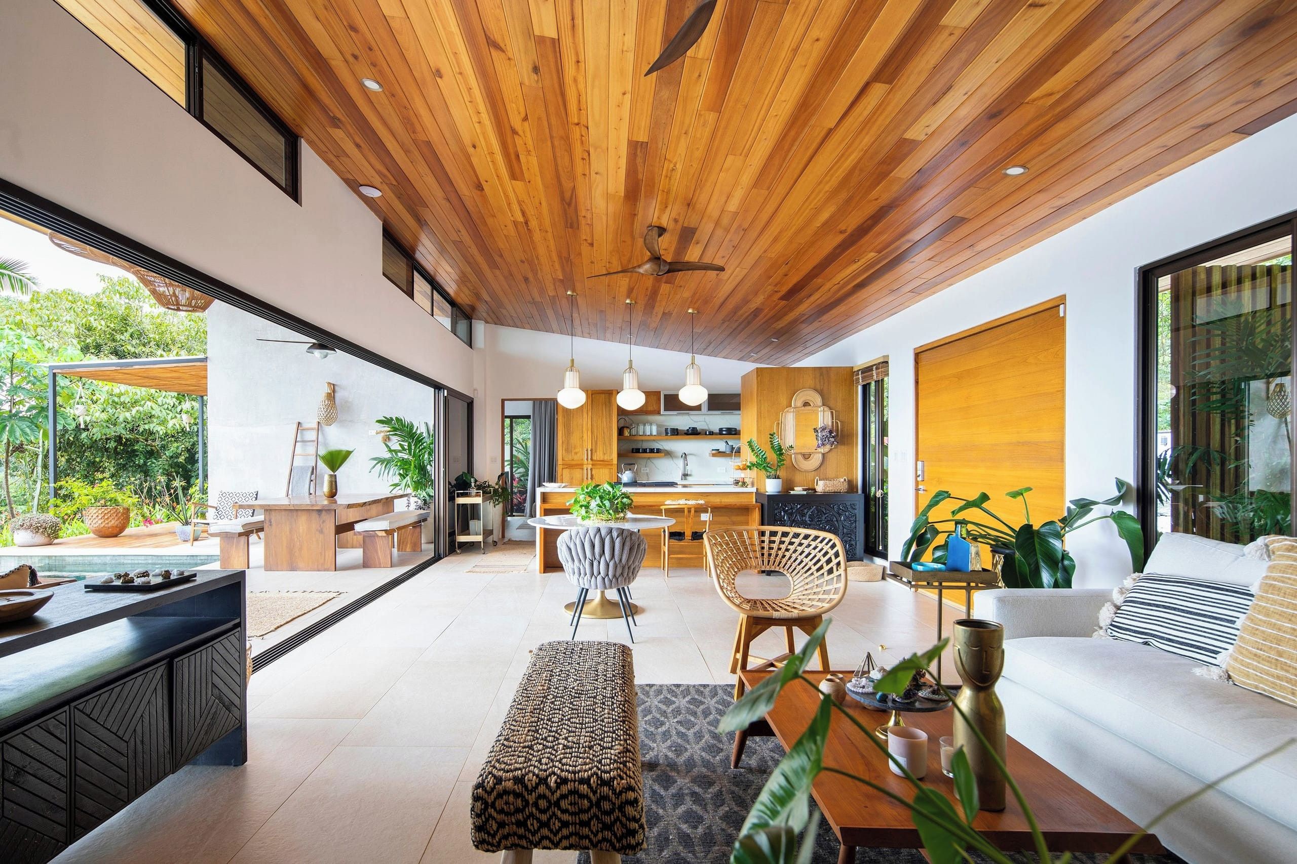 A modern interior with a wooden ceiling, a dining area, a kitchen, and a living room with a sofa and plants.