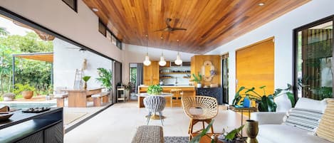 A modern interior with a wooden ceiling, a dining area, a kitchen, and a living room with a sofa and plants.