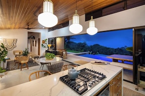 A modern kitchen with a central island, a dining area with wicker furniture, and a large window overlooking a pool. - Hidden Villa Oasis: Infinity Pool & Ocean Views near Dominical (Savegre de Aguirre)