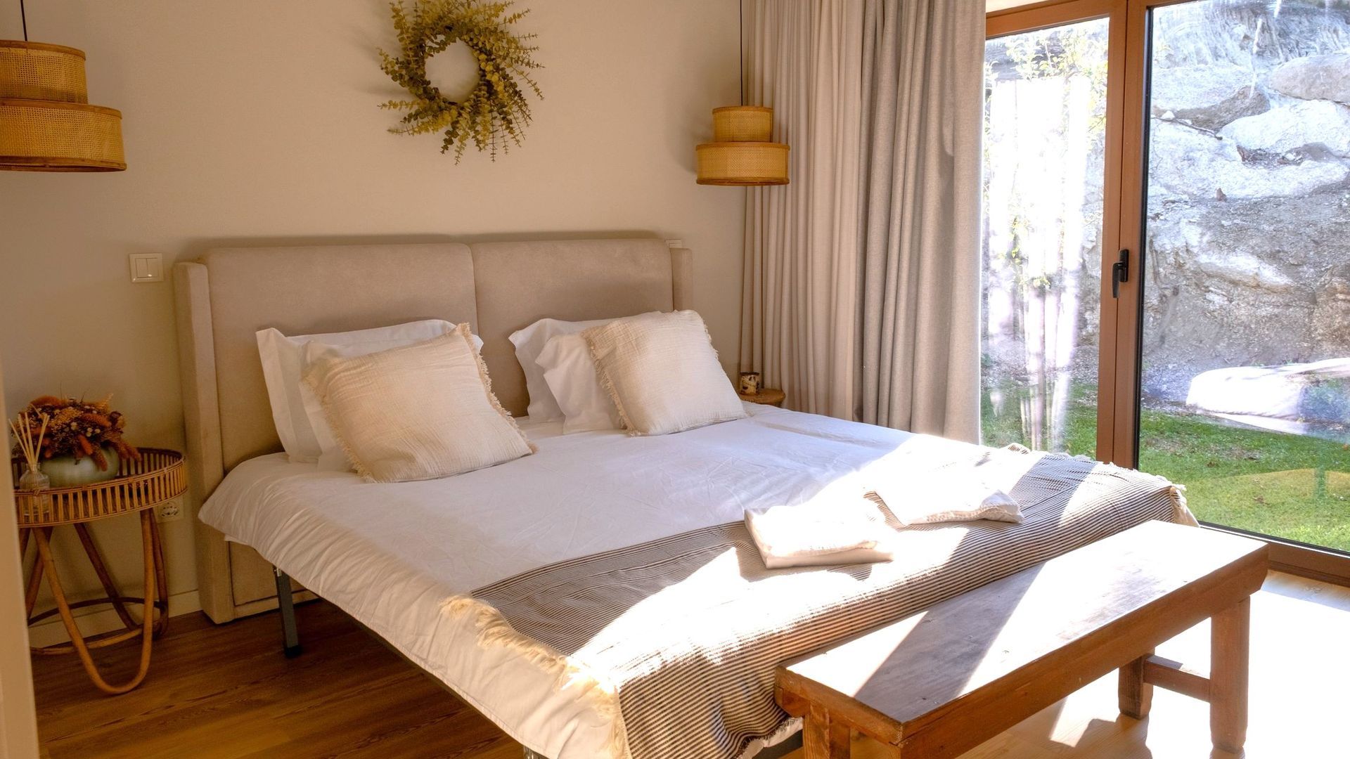 A bedroom with a bed, a bench, a wall-mounted lamp, a side table with flowers, and a view of a rocky outdoor area.