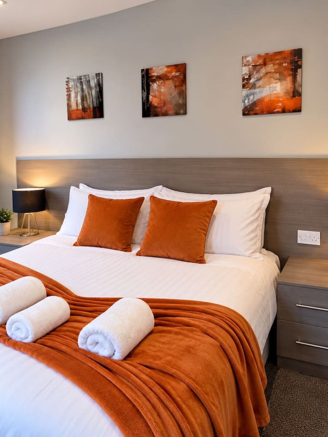 A hotel room with a bed, two pillows, a blanket, a nightstand, a lamp, and three abstract paintings on the wall.