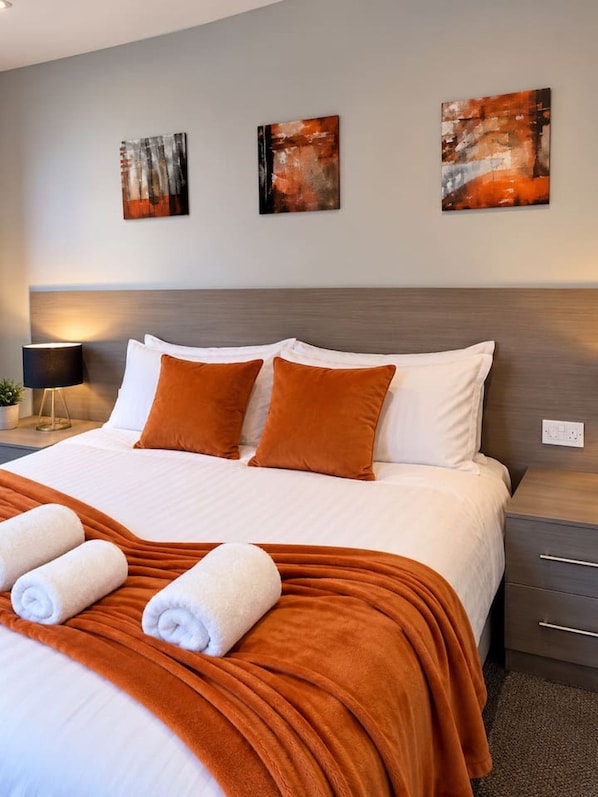 Una habitació d'hotel amb un llit, dos coixins, una manta, una tauleta de nit, una làmpada i tres quadres abstractes a la paret.