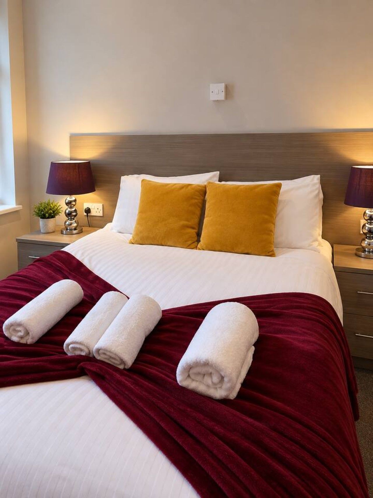 Una cama bien hecha con almohadas blancas y amarillas, una manta roja y toallas enrolladas.