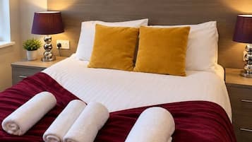 Una cama bien hecha con almohadas blancas y amarillas, una manta roja y toallas enrolladas.