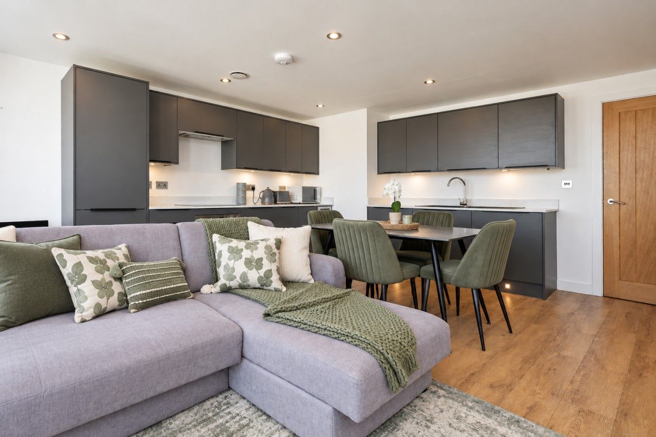A modern living room with a grey sofa, a dining area with green chairs, and a kitchen with dark cabinets.
