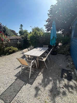 A patio with a table and chairs, a blue umbrella, and a gravel ground. - La Noisette & Le Jardin de Sasha (sleeps 6) (Équemauville)
