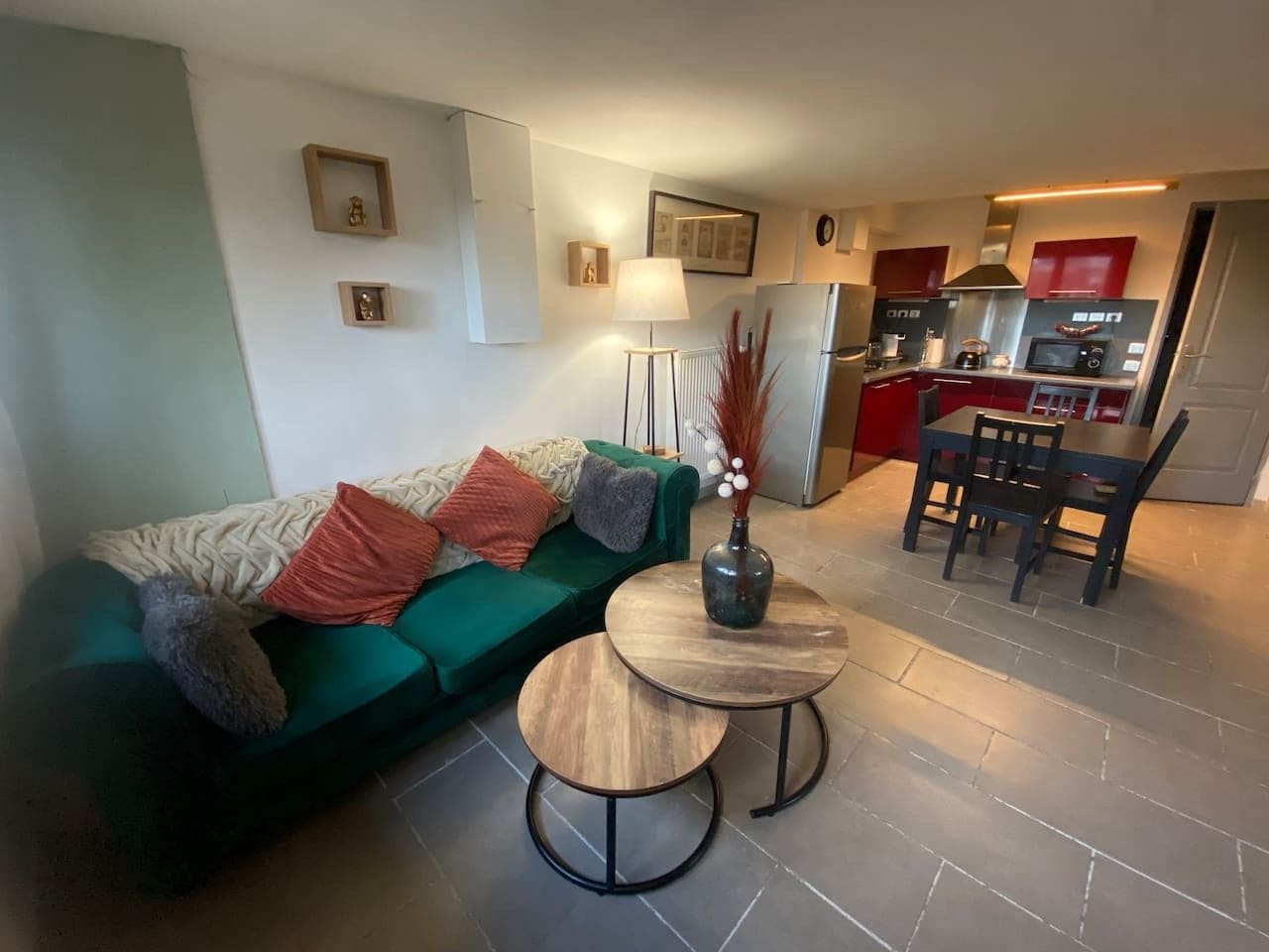 A living room with a sofa, two side tables, a lamp, and a vase. There is a kitchen area with a refrigerator, microwave, and dining table with chairs.