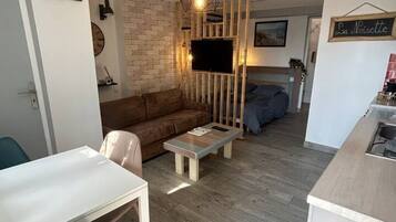 A compact living space with a sofa, a coffee table, a TV mounted on a wooden partition, and a small kitchenette area.