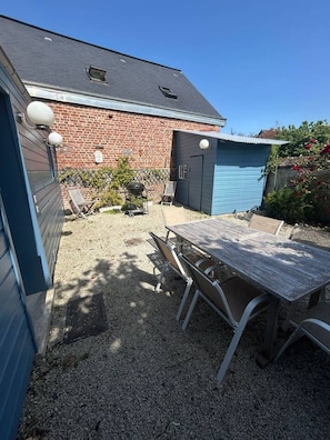 Um quintal com um pátio de cascalho, uma mesa de madeira e cadeiras. Há um prédio de tijolos com uma porta de garagem azul e um pequeno galpão.