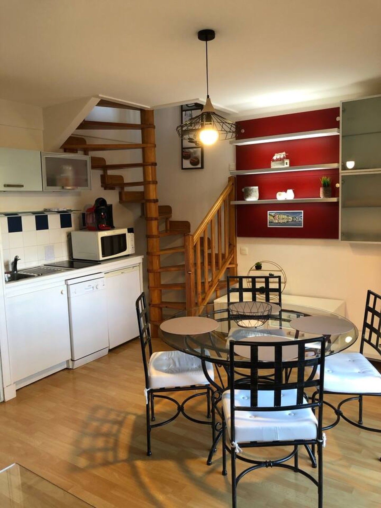 A compact kitchen with white appliances, a dining area with a round glass table and black chairs, and a red shelving unit.
