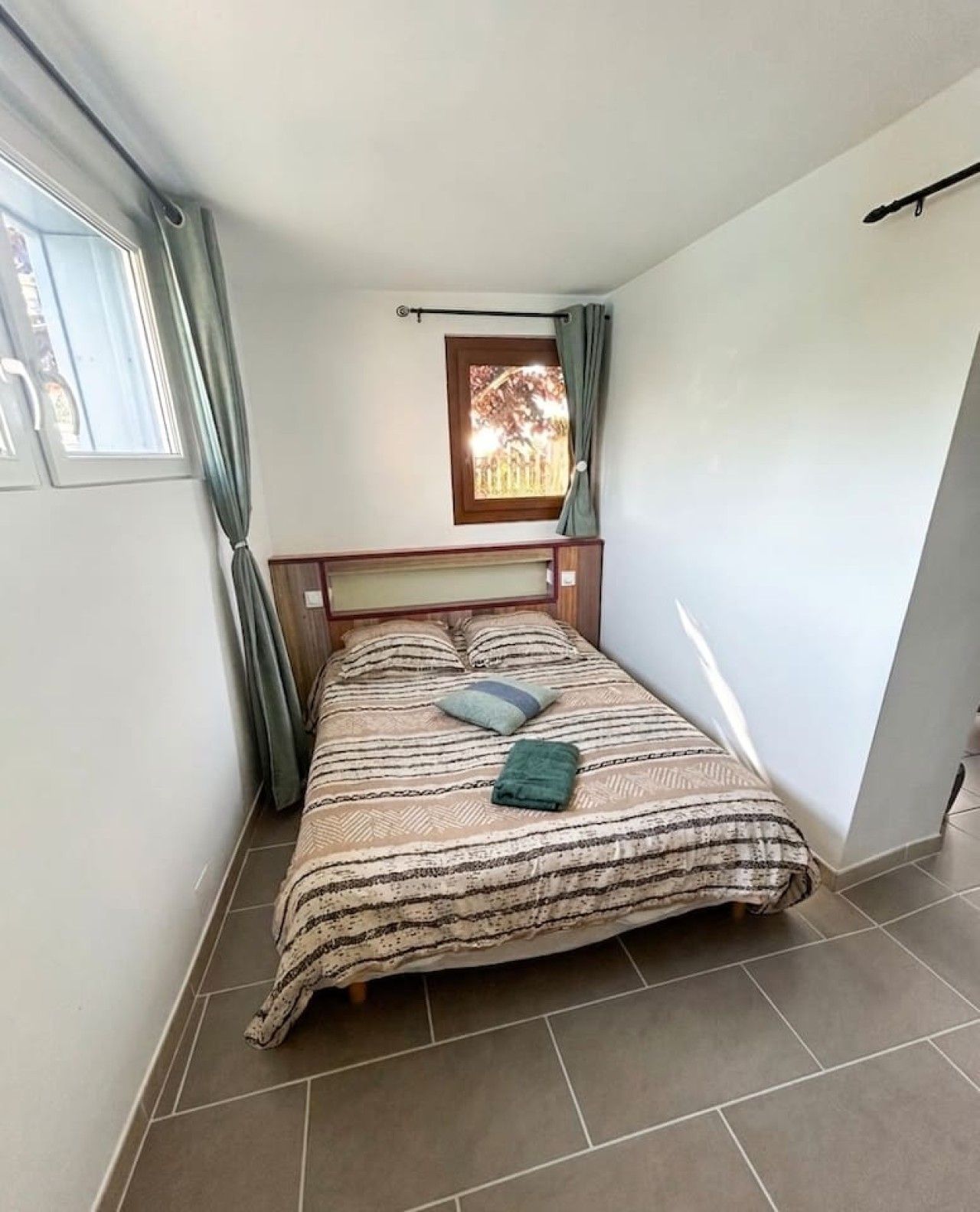 Un lit simple avec une couvre-lit à motifs, un oreiller et une serviette pliée. Il y a une fenêtre avec des rideaux, un cadre photo accroché au mur et un radiateur.