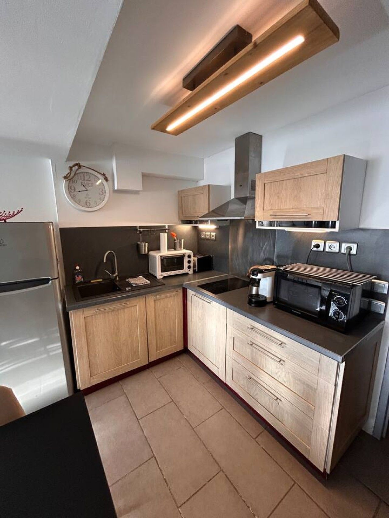 A compact kitchen with wooden cabinets, a built-in microwave, a toaster, and a clock on the wall.