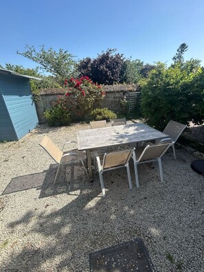 Un patio al aire libre con mesa y sillas, rodeado de grava, un cobertizo azul y diversas plantas.
