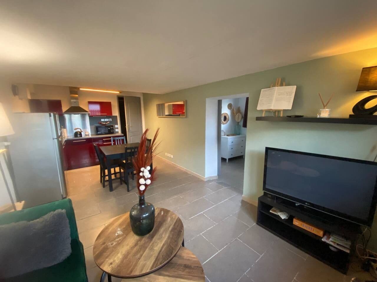 A modern living room with a flat-screen TV, a dining area with a table and chairs, a kitchen in the background, and a laundry area with a washing machine.
