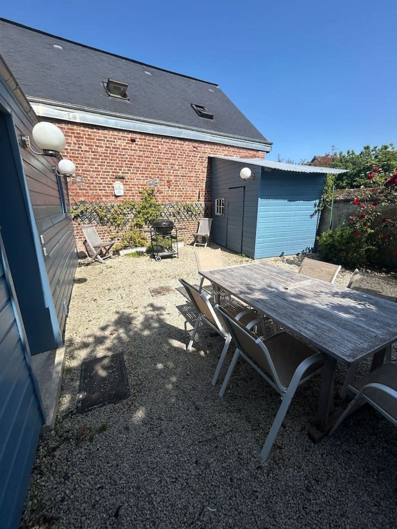 A backyard with a gravel patio, a wooden table with chairs, a barbecue grill, and a blue shed.