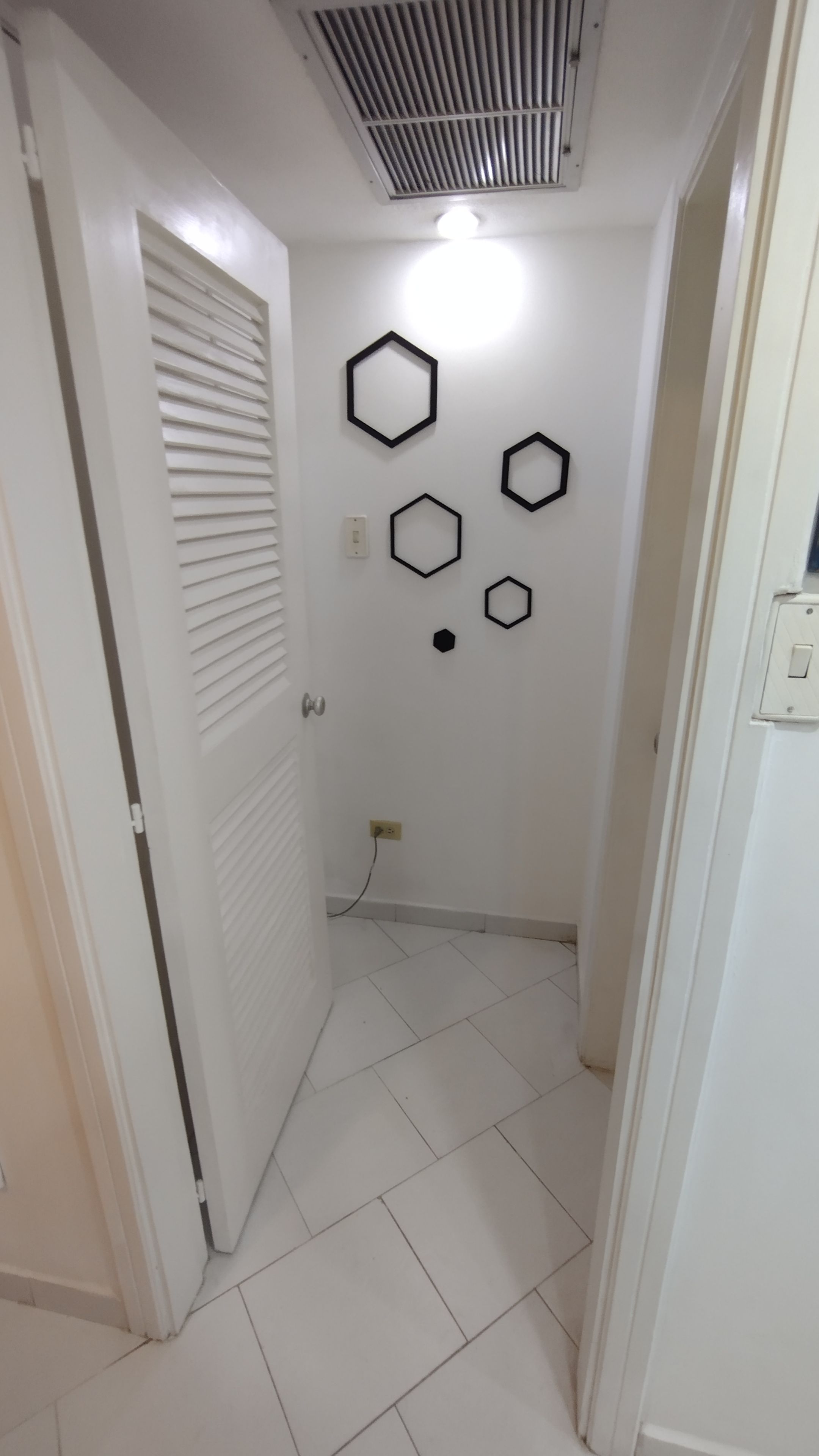 A narrow hallway with white tiled flooring, a door with a louvered window, and a wall with hexagonal wall art.