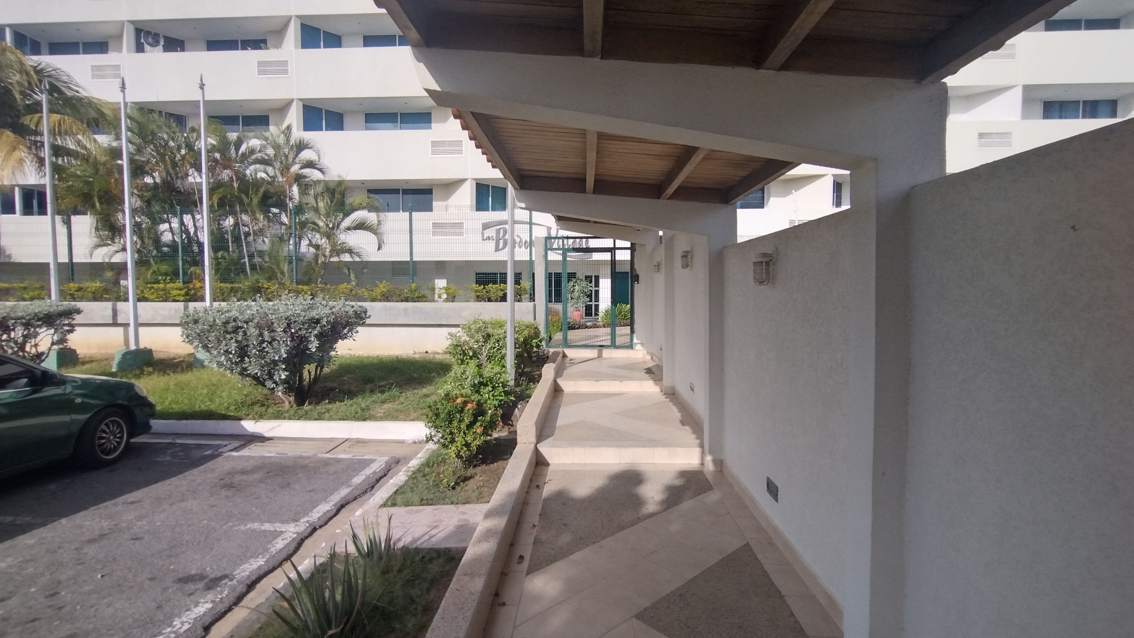 A covered walkway leading to a building with a gated entrance, surrounded by greenery and a parked car.