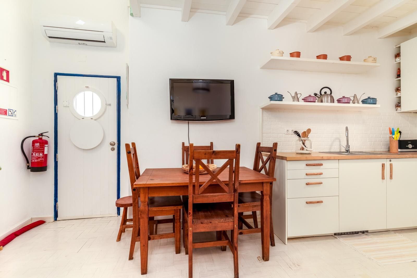 A compact kitchen with white cabinets, a wooden dining table, and a flat-screen TV mounted above it.