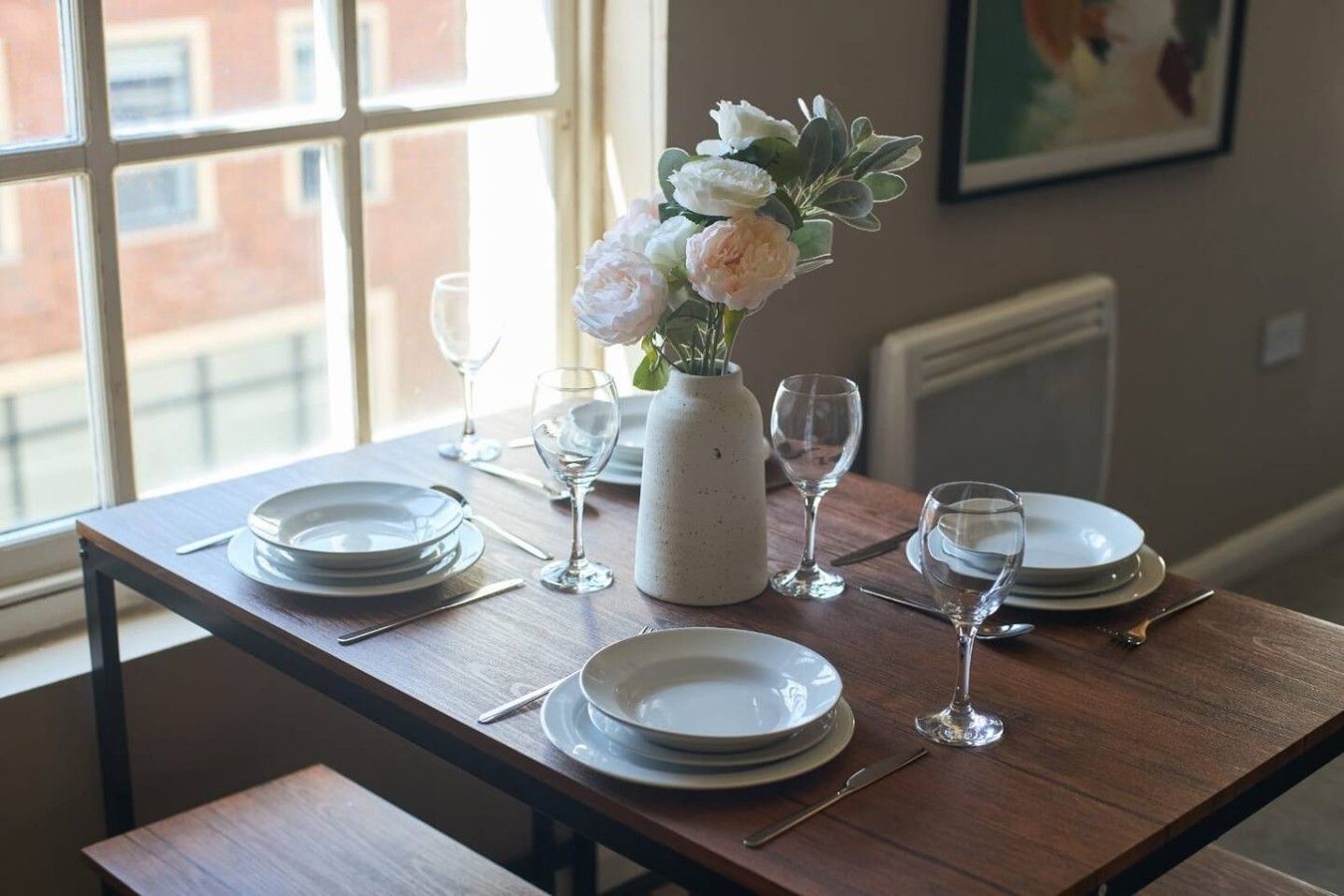 A dining table set for four with plates, cutlery, and wine glasses, a vase with flowers, and a window with a view of buildings.
