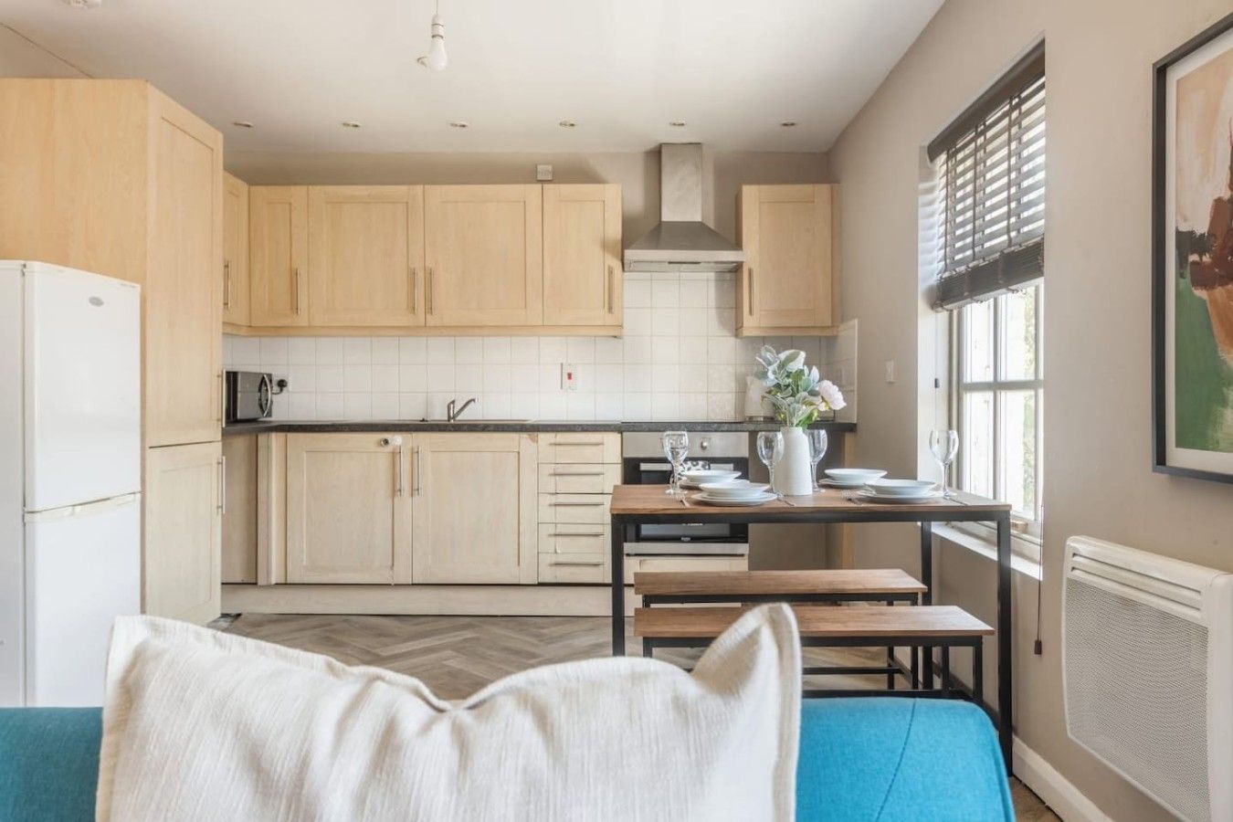 A modern kitchen with light wood cabinets, a dining table set with plates and glasses, and a window with blinds.