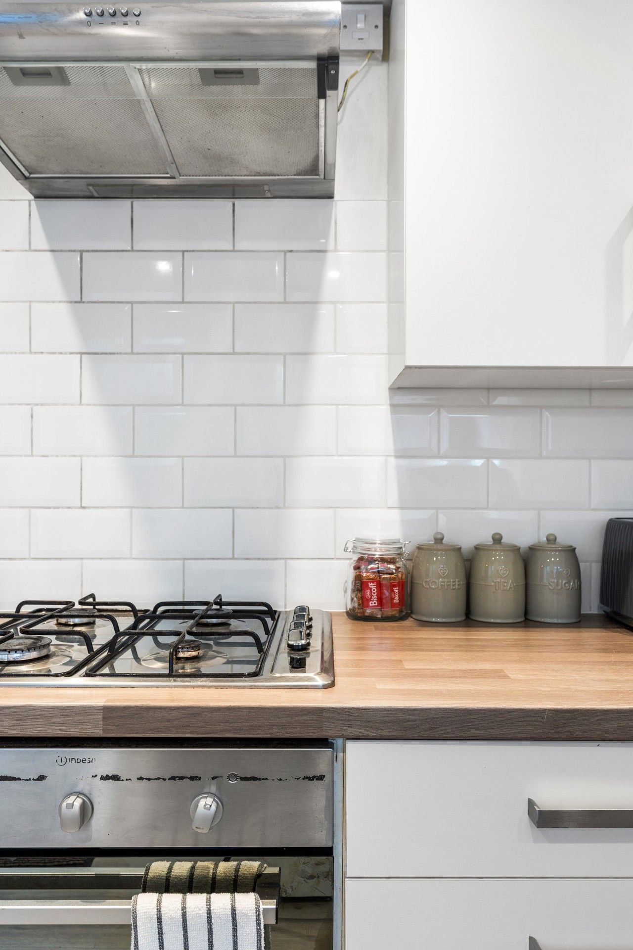 A kitchen with a gas stove, a stainless steel range hood, white tiled backsplash, and a wooden countertop with jars labeled 'Coffee,' 'Tea,' and 'Sugar.'
