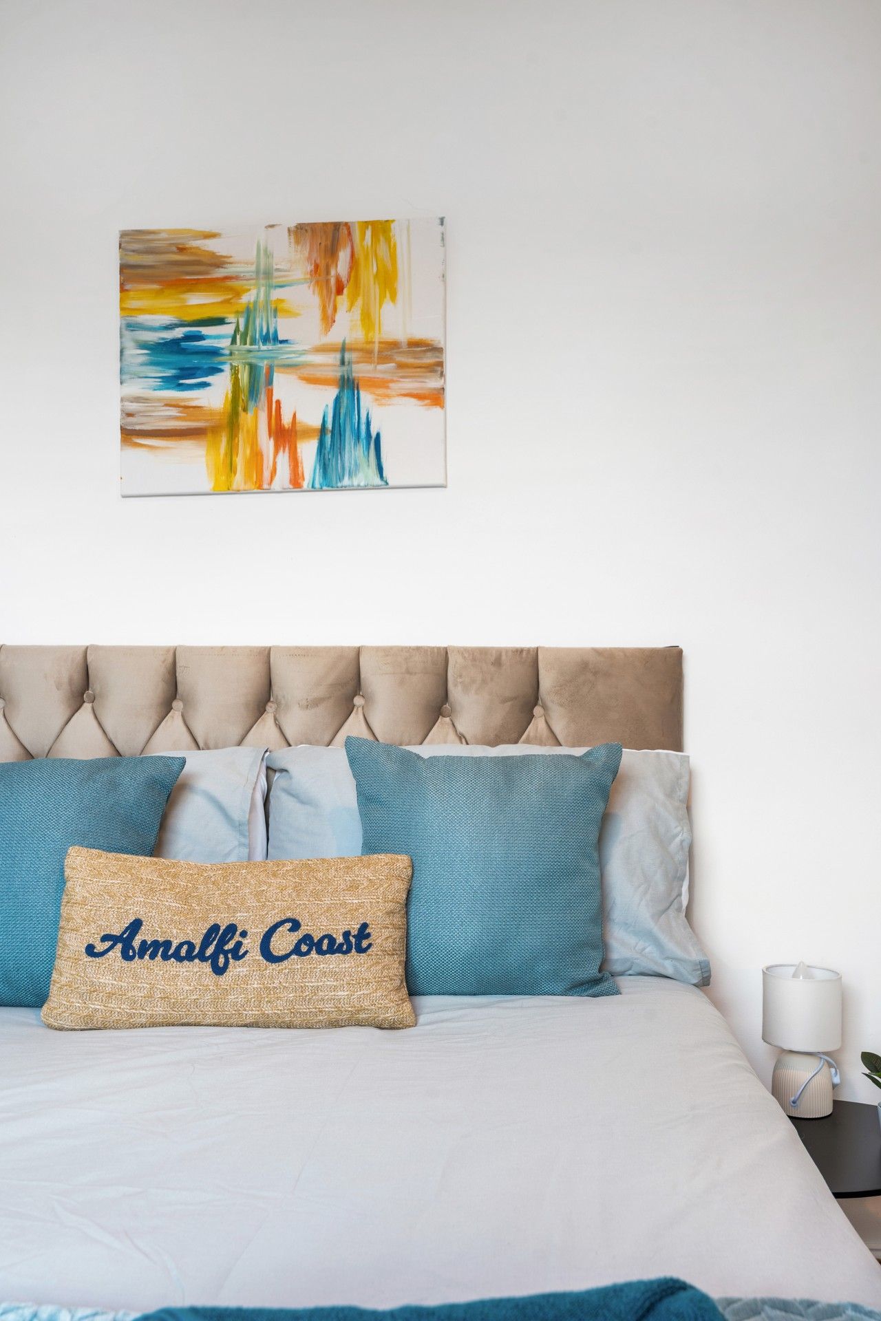 A bedroom with a tufted headboard, a bed with multiple pillows, a wall-mounted abstract painting, and a bedside table with a lamp.