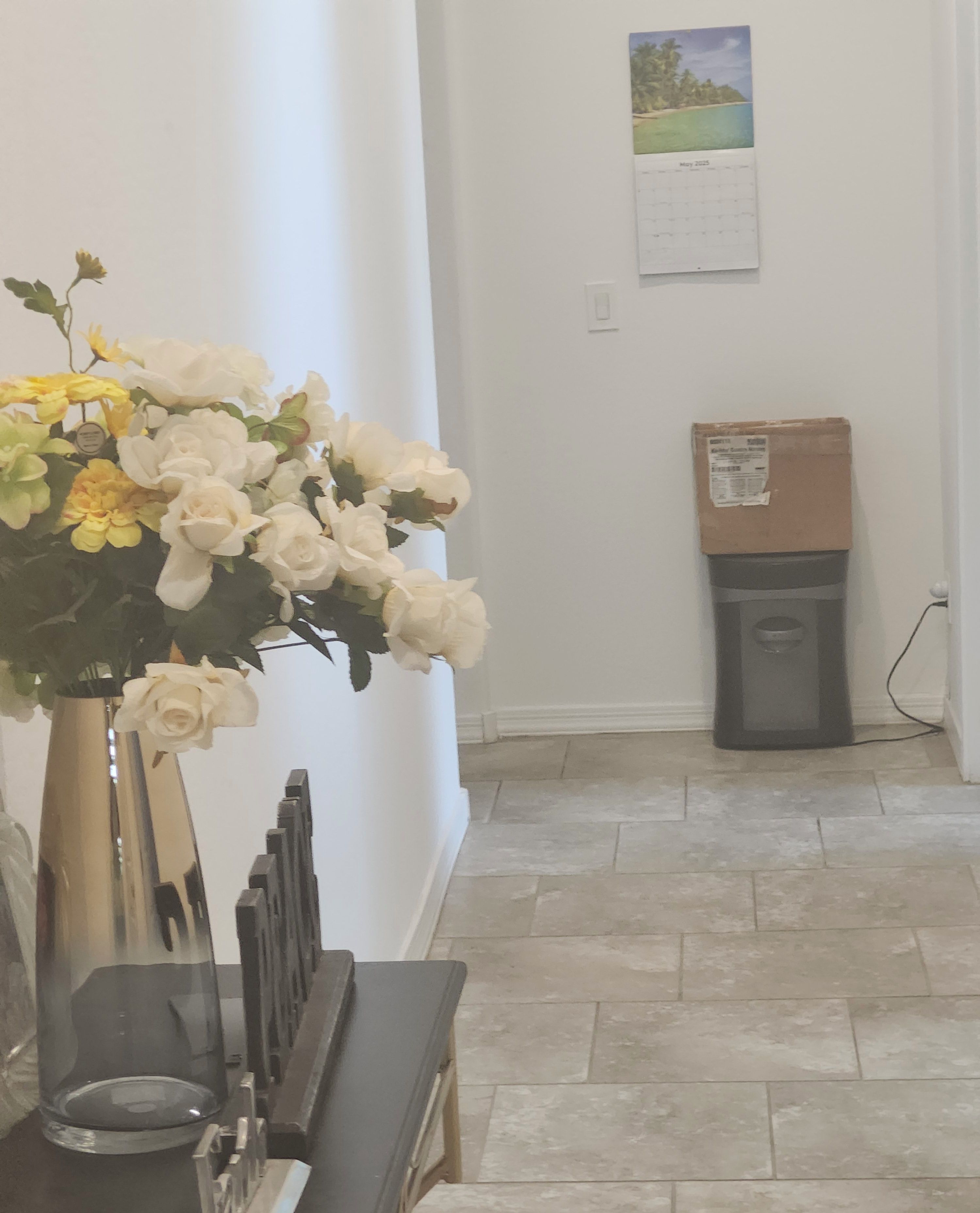 A hallway with a large white vase of flowers, a calendar on the wall, and a cardboard box on the floor.