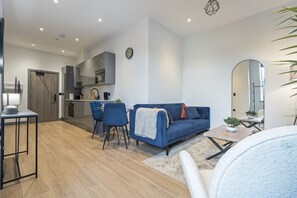 A modern living room with a blue sofa, a dining table, and chairs. There is a kitchen area in the background with a clock on the wall. - N. Finchley Retreat | 2 Bed | 2 Bath | Sleeps 5 (North Finchley)