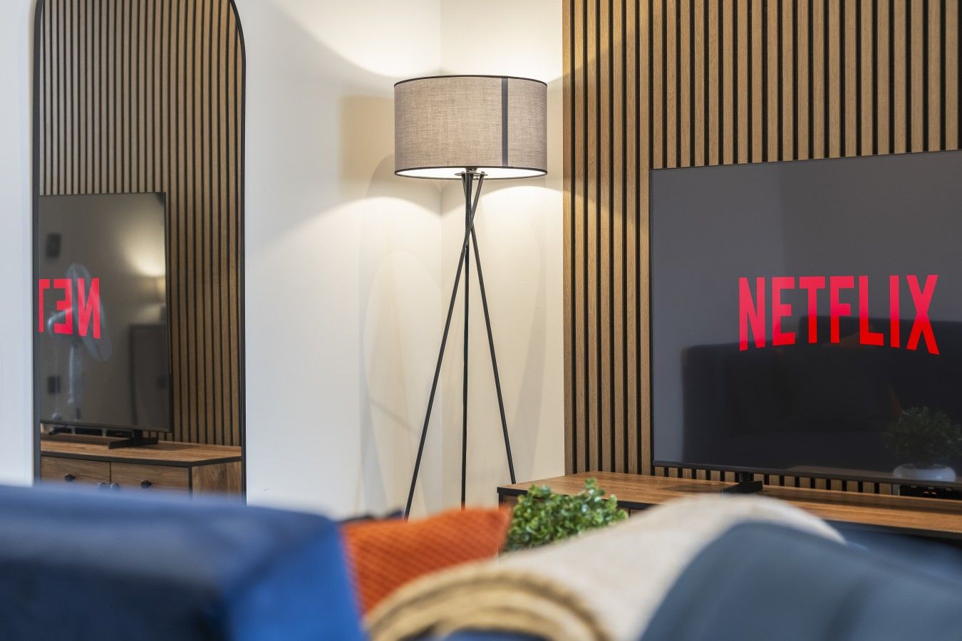 Moderni olohuone, jossa on televisio, jonka näytöllä näkyy punaisilla kirjaimilla teksti »NETFLIX», lattiavalaisin ja puupaneloitu seinä.