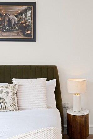 Quarto com cama, almofadas, mesa de cabeceira, abajur e quadro na parede.