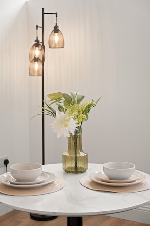 Mesa de jantar redonda com duas tigelas e pires brancos, um vaso dourado com flores brancas e um abajur com duas cúpulas.