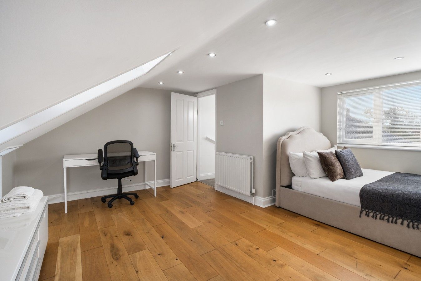 Une pièce avec un plafond en pente, un bureau blanc avec un fauteuil de bureau noir, un radiateur et un lit avec des oreillers.