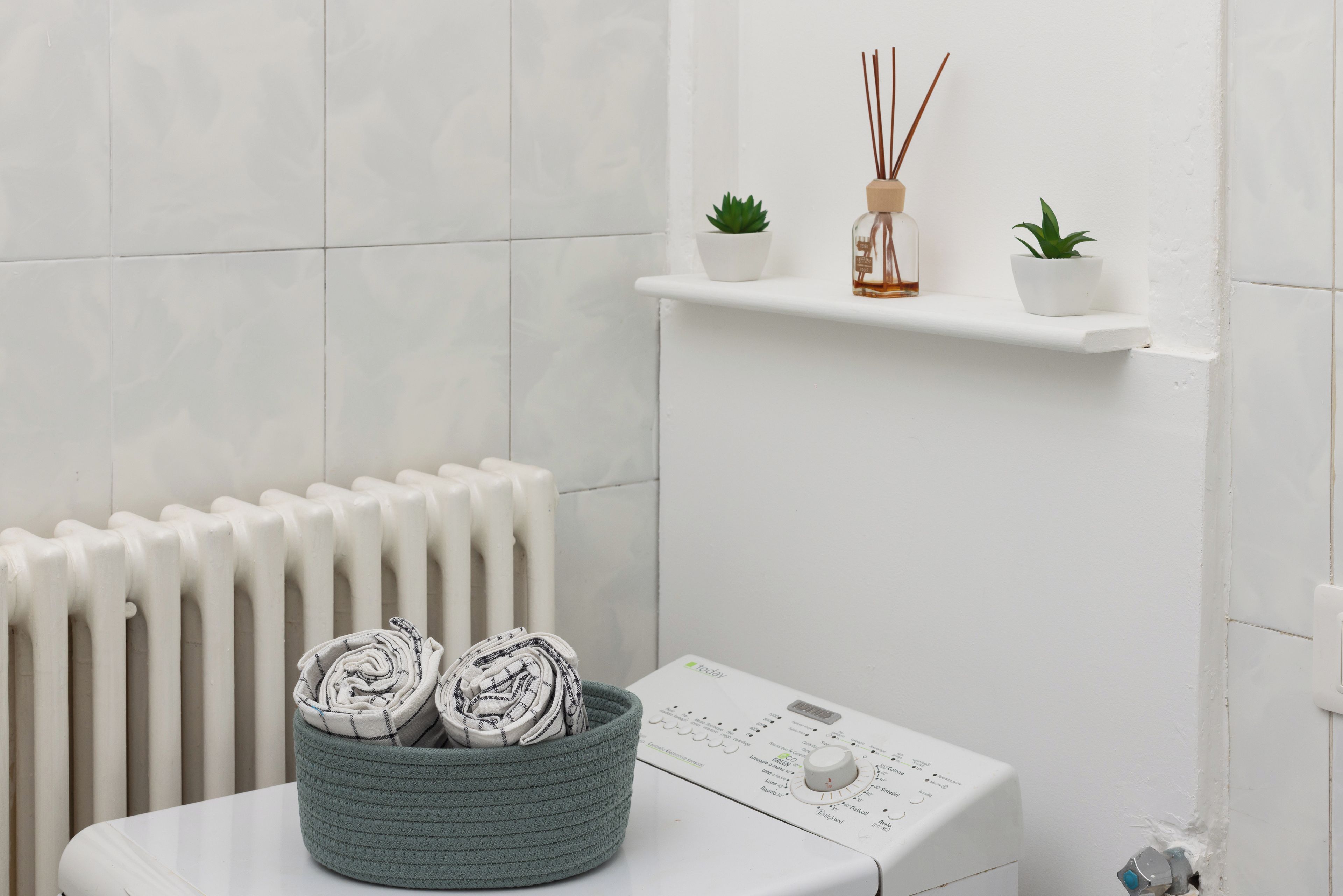 Ein Badezimmer mit einem weißen Heizkörper, einer weißen Waschmaschine, einem grünen Korb mit zusammengerollten Handtüchern und zwei kleinen, in Töpfen stehenden Pflanzen auf einem weißen Regal.