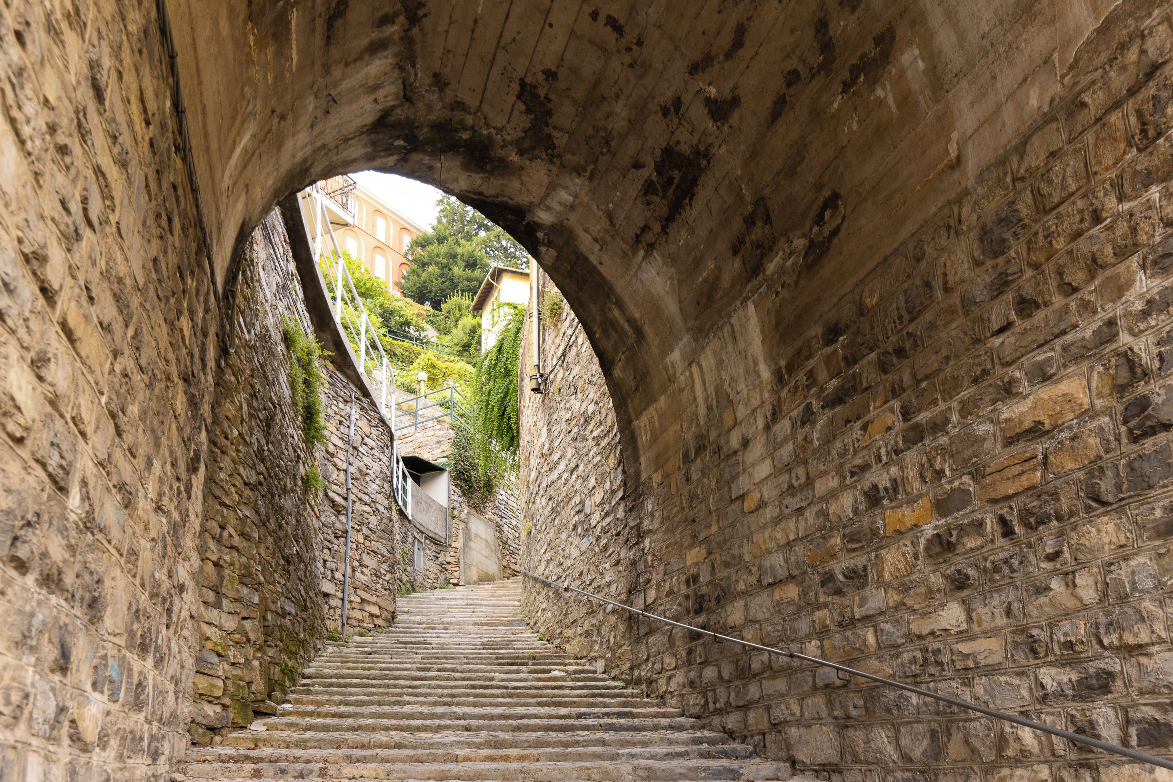 Un escalier en pierre mène à travers un tunnel, d’où l’on aperçoit un bâtiment et de la verdure à l’extérieur.